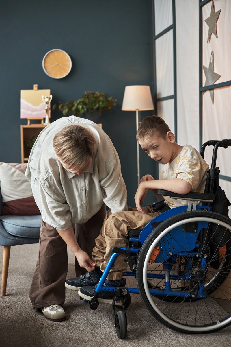 A caregiver bending down and helping a boy with disability in wheelchair inside cozy home. Caring interaction between adult and child fostering support and encouragement