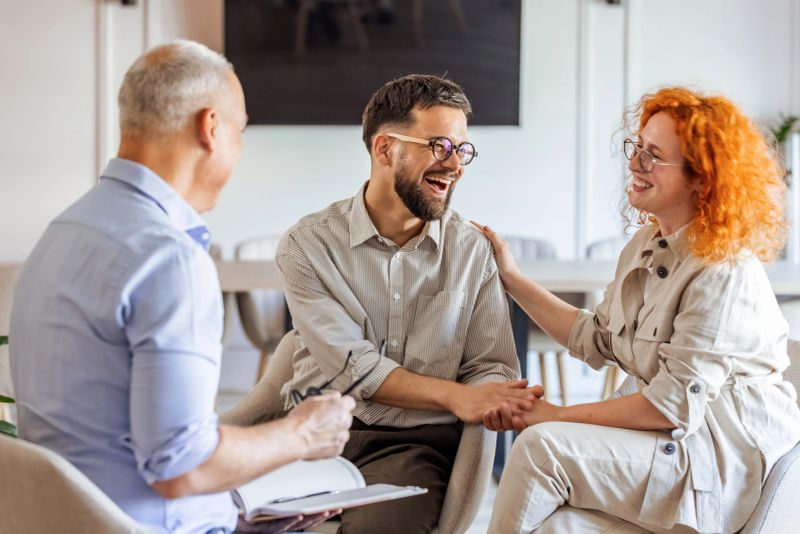 A joyful couple engages warmly with a counselor during a therapy session, showcasing a supportive and loving interaction. A bright office setting enhances the feeling of warmth and comfort in the environment.