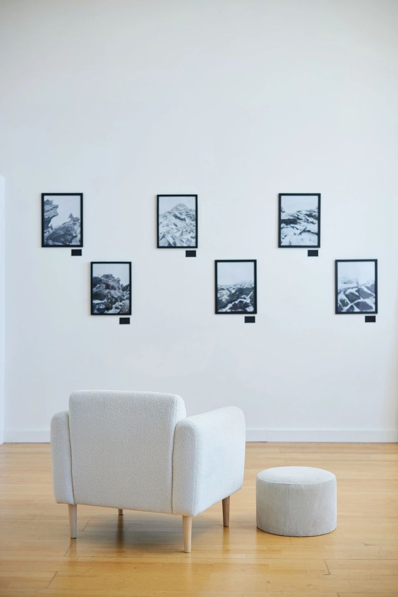 Art exhibition displaying black and white photographs in minimalist setting with white sofa and round ottoman in foreground on wooden floor creating serene atmosphere