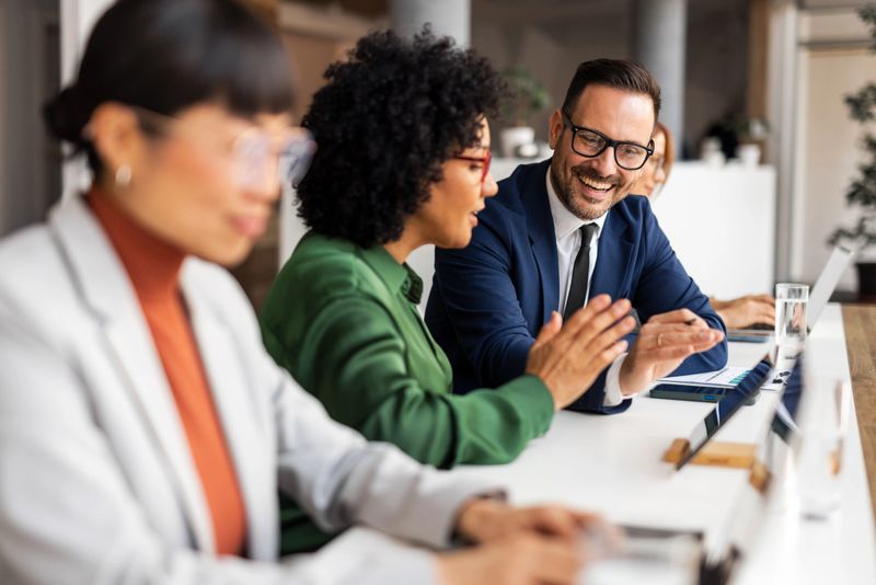 A group of coworkers discussing ideas in an office. They are smiling, interacting, and sharing perspectives, showcasing teamwork and professionalism. Laptops and documents emphasize a modern corporate setting.