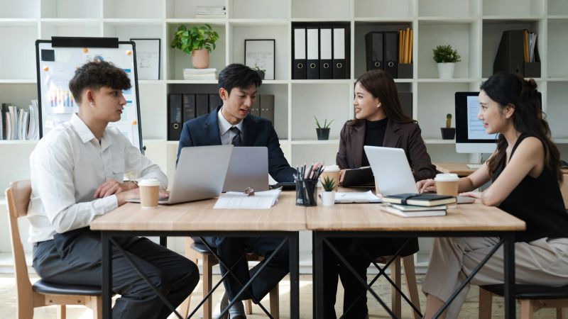 A diverse group of young professionals engaged in a business meeting around a conference table, using laptops and documents in a bright, modern office space.