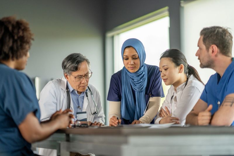 A diverse team of healthcare professionals gathers around a table discussing important medical cases in a professional setting, showcasing collaboration, diversity, and dedication in the medical field during a team consultation.