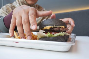 Person reaching for fries next to a black bun cheeseburger on a tray.