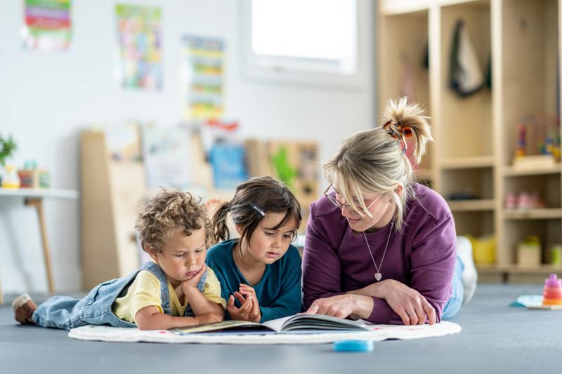 A teacher and two children engage in a storybook activity on the floor inside a classroom, fostering education, interaction, and creativity in a supportive and playful environment.