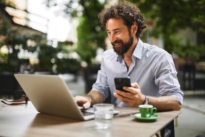 Man working on laptop and holding phone outdoors.