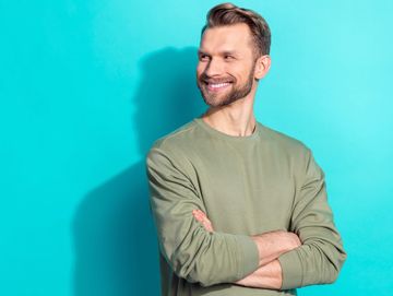 Smiling man in green sweater standing against a blue wall.