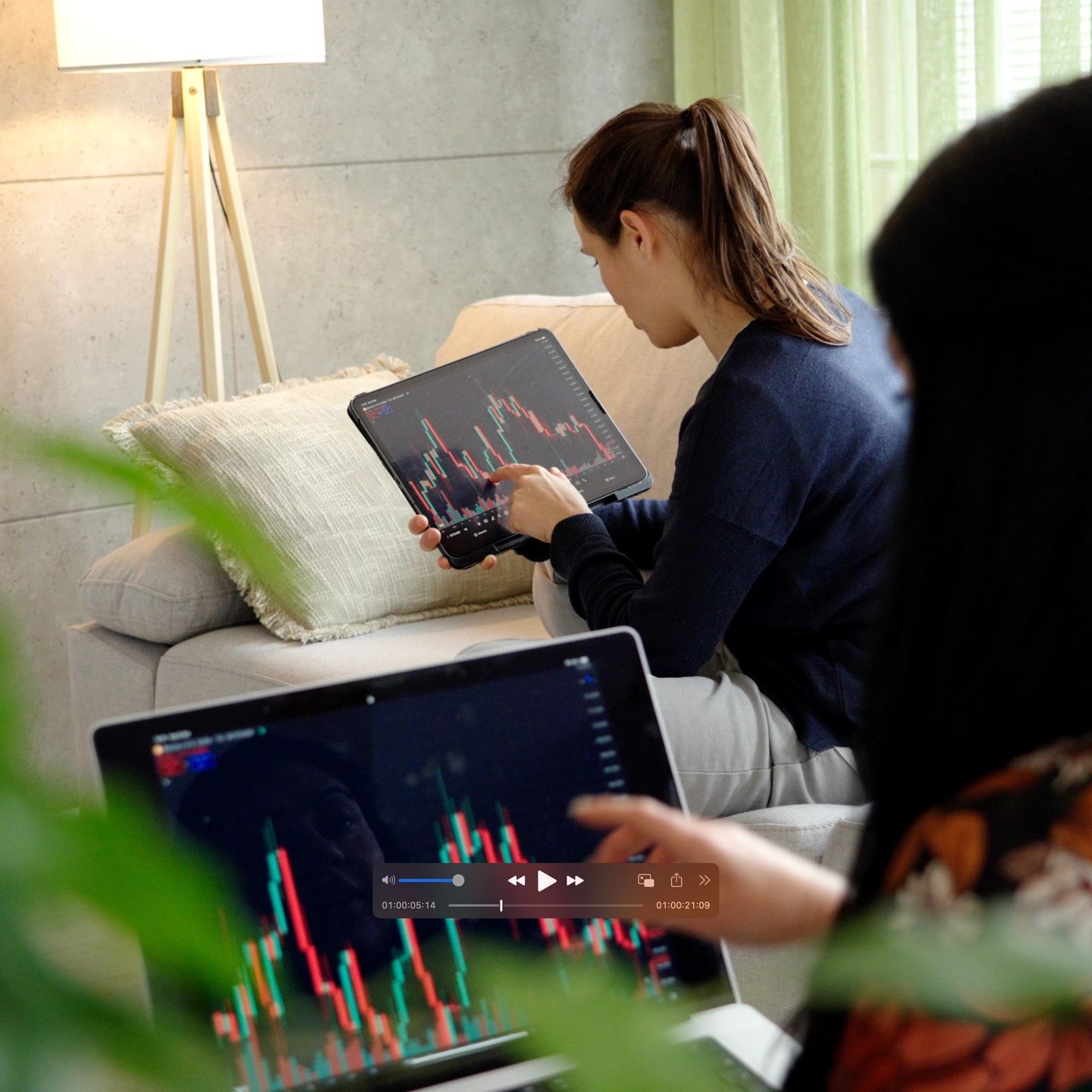 Two women analyzing stock charts on digital devices in a cozy living room.