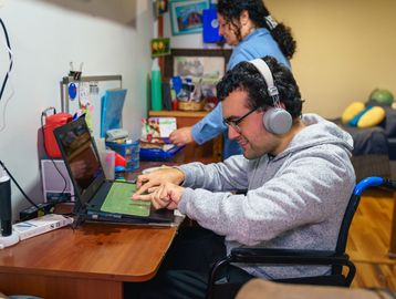 A man in a wheelchair using a laptop with another person in the background.