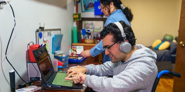 A man in a wheelchair using a laptop with headphones on, while a woman stands in the background.