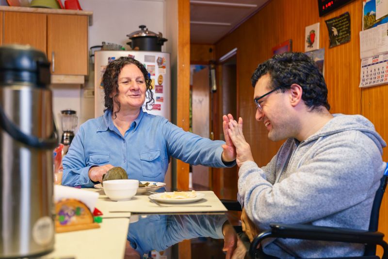 Heartwarming moment between a latin american woman and a young man with cerebral palsy, sharing a high-five during a meal at home