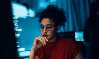 Young woman in glasses focused on a computer screen in a dimly lit room.