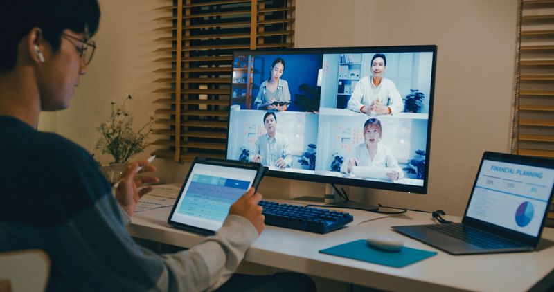 Closeup of young Asian man using digital tablet and laptop for video conference with colleagues at night from his home office, showcasing remote work and teamwork. Remotely and finance concept.