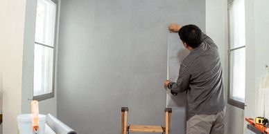 Man applying gray wallpaper in a room with two windows.