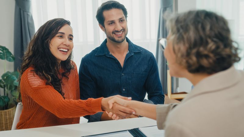 Financial advisor using tablet discussing about life insurance domestic finances and handshake agreement with young couple in living room at home. Financial planning and modern lifestyles concept.