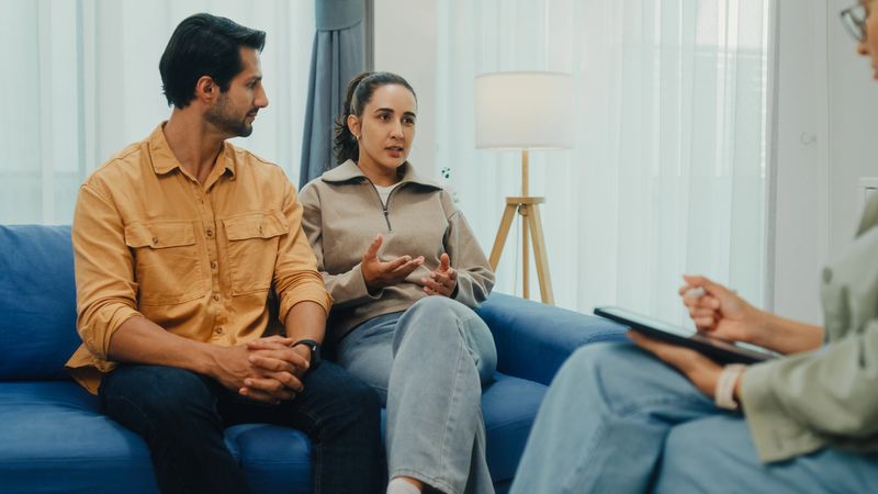 Young couple in serious discussion with mental health professional during marriage counseling session sit on sofa in living room at home. Therapy session and mental health concept.