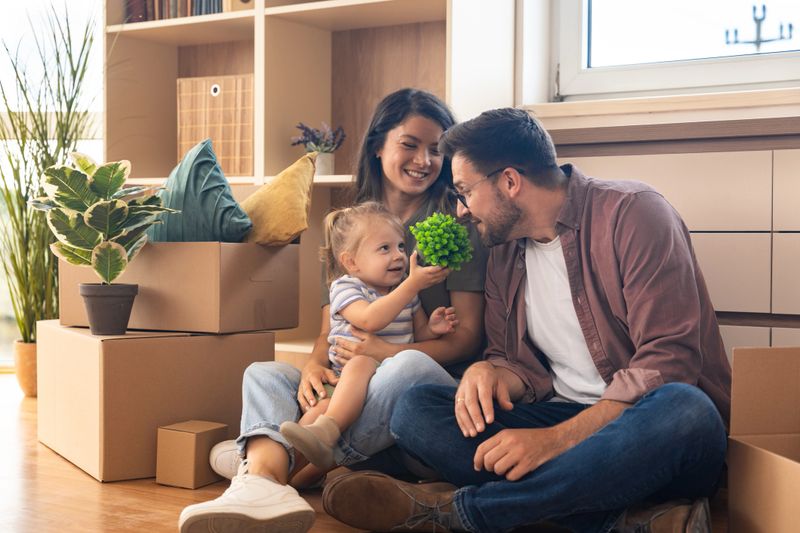 Happy family moving into new home. Young man and woman, parents of small baby girl, having fun with their child, while unpacking cardboard boxes starting their new independent life at new apartment.