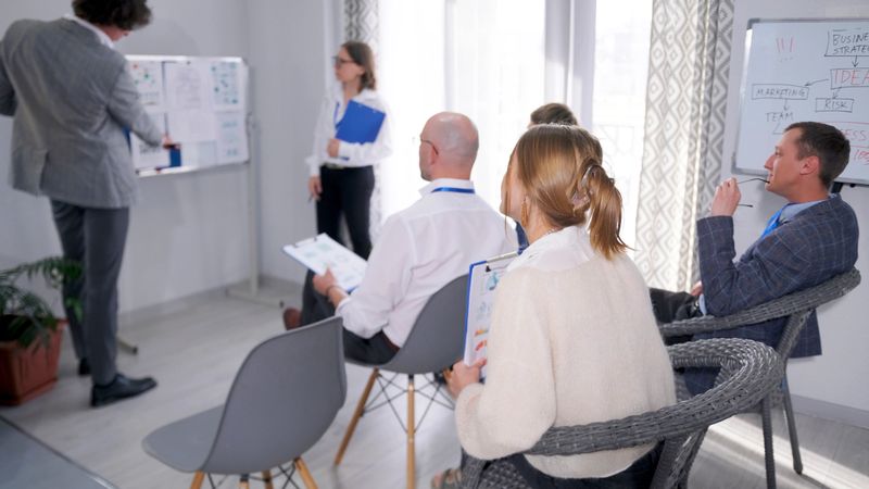 The company's staff sits on chairs in the room and listens to the presentation. Mentoring. Team building. Morning briefing. The presentation is conducted on a white board by a man and a woman