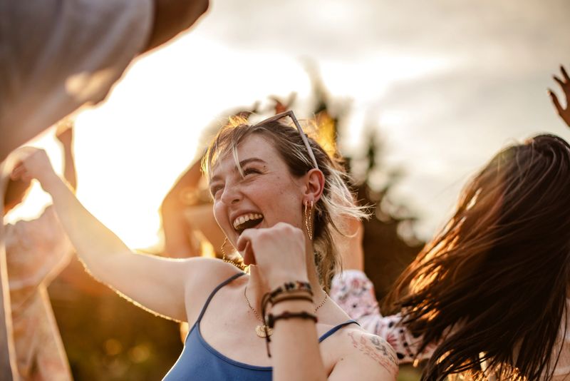 A lively crowd of young adults dances freely at an outdoor concert, celebrating music with arms lifted in excitement. The sunset casts a warm glow over joyful friends enjoying the moment.