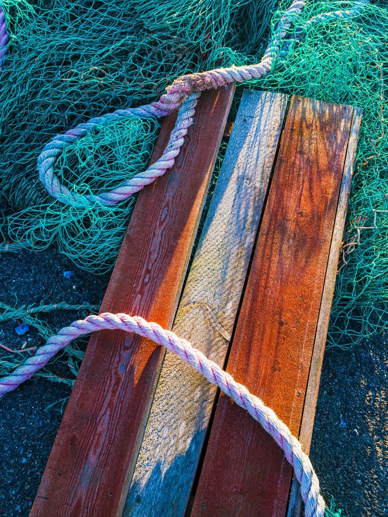 A collection of aged wooden planks lies on the ground, entwined with vibrant green fishing nets and twined ropes, illuminated by the afternoon sunlight at a seaside area.