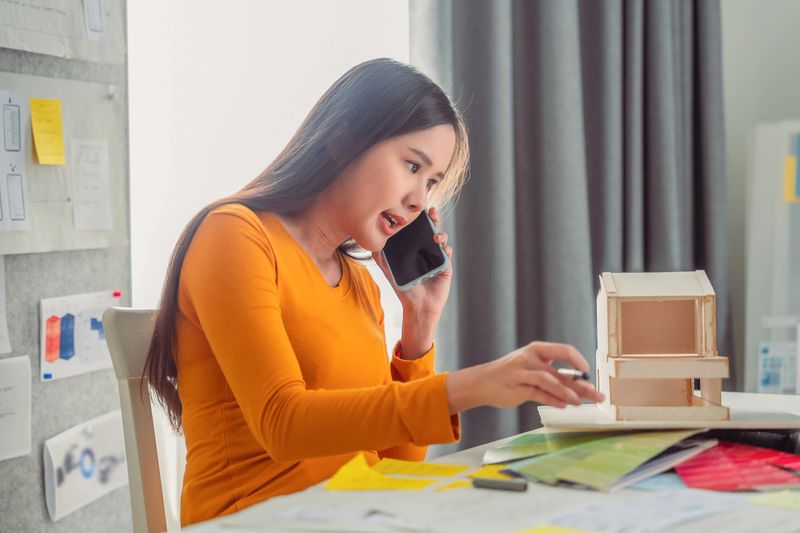 A focused Asian female architect smiles gently while working from her home office, using her smartphone to review blueprints and coordinate a new house design project. Surrounded by drafting tools and modern decor, she balances creativity and technology in her remote architectural workspace, showcasing professional design excellence and home productivity.