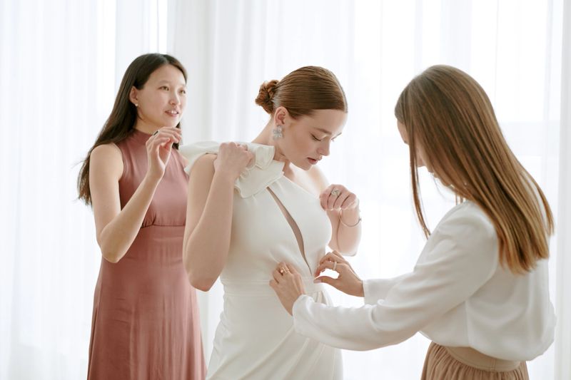 Caucasian young adult woman standing while two young adult women assisting with wedding dress fitting, preparing for wedding celebration in bright room with sheer curtains