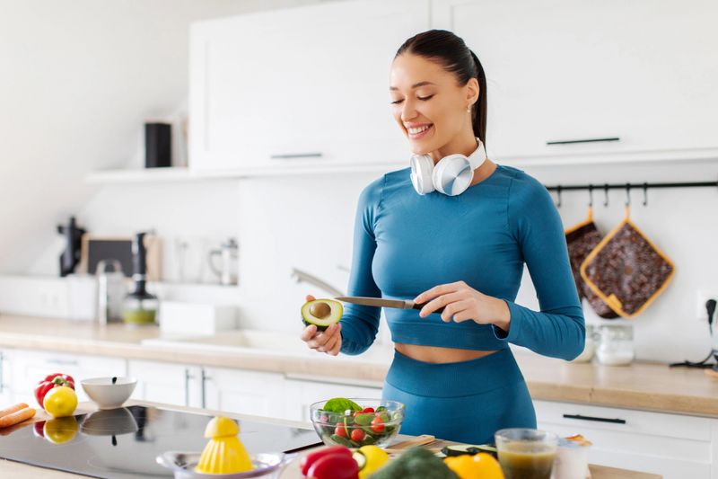 Beautiful fit lady making healthy salad, peeling avocado and cooking dinner in the kitchen at home, free space. Woman wearing sportswear and headphones on neck