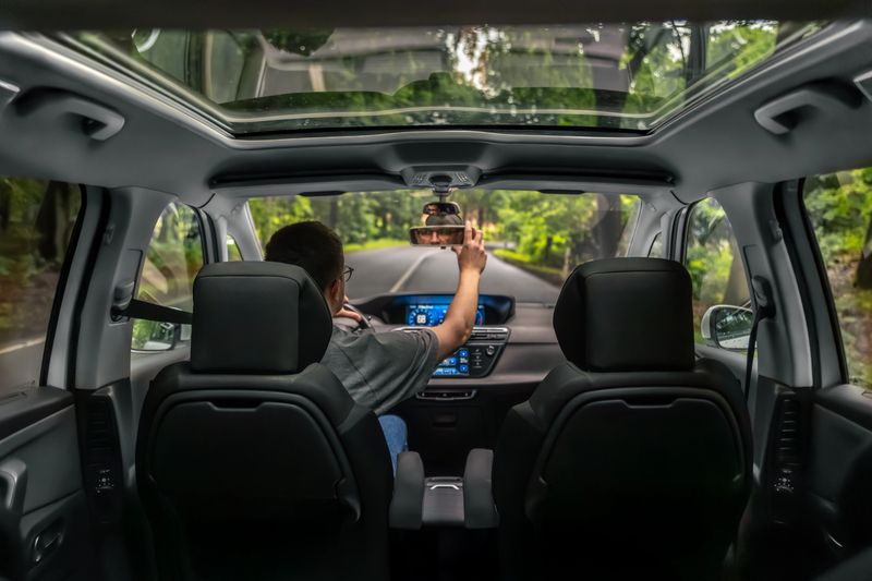 Man driving a car adjusts the rearview mirror while traveling along a forest road. The modern interior features a panoramic roof and digital dashboard.