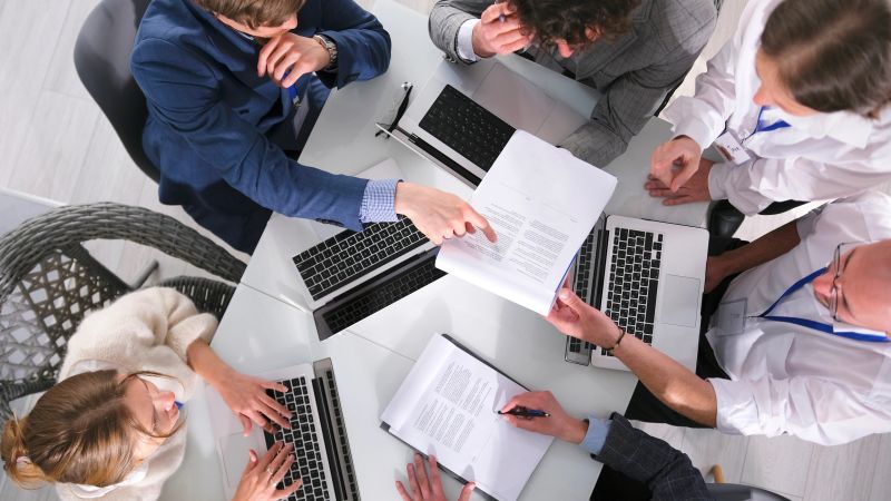 The company's lawyer advises to draw the boss's attention to the important clause in the agreement, top view. A group of colleagues sits at a table with laptops during a meeting.