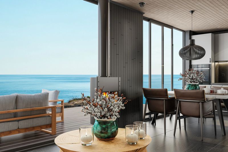 Sophisticated open-plan dining and kitchen space featuring contemporary Scandinavian design with stunning floor-to-ceiling glass windows offering panoramic views of the sea. The interior includes a sleek white kitchen, matte black tile flooring, a central modern dining table with leather chairs, and a cozy wood-burning fireplace. A statement black pendant light and decorative vase add character, while the warm wood ceiling and outdoor seating area create a seamless transition between indoor comfort and coastal living. Ideal for showcasing architecture, interior design, real estate, and luxury lifestyle concepts.