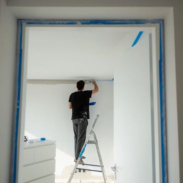 Person painting a white wall using a ladder inside a room.