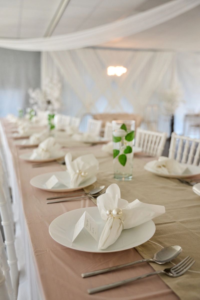 Elegant wedding reception table is set with white napkins, pearl rings, and greenery under a white canopy