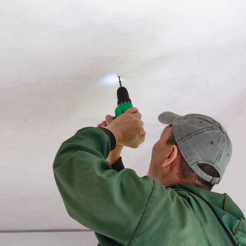 Worker wearing green jacket and grey cap uses cordless electric screwdriver to install or repair ceiling indoors, bottom view.