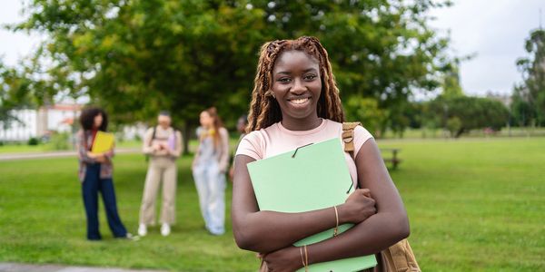 Smiling student holding a folder outdoors on campus with friends in the background.