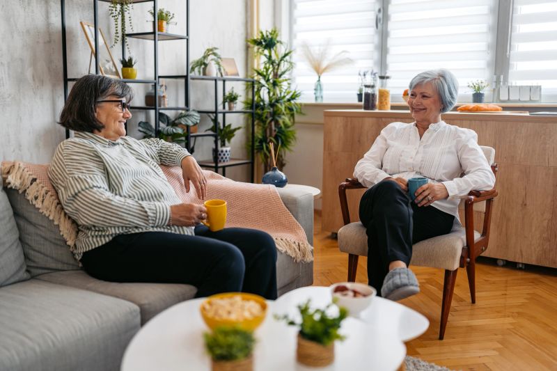 Two senior female friends drinking coffee and talking on the sofa in the living room.