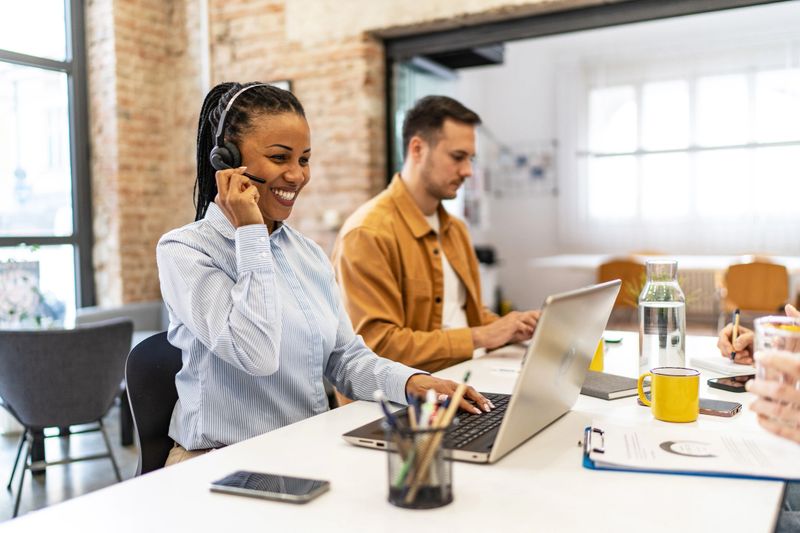 Cheerful customer service representative wearing a headset smiles while assisting clients on her laptop in a bright, modern office, her colleagues working diligently beside her