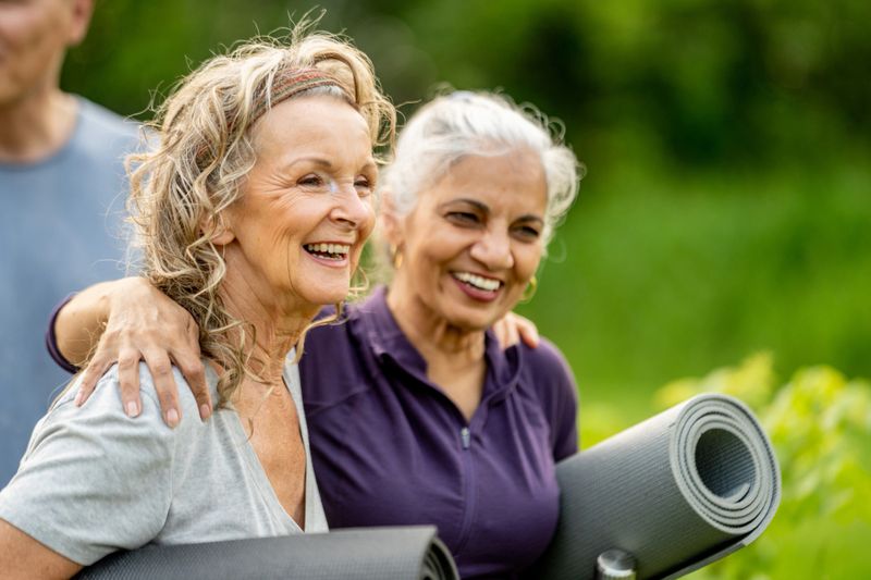 Two older women share a joyful moment outdoors, holding yoga mats and smiling warmly. The setting highlights health, companionship, and the enjoyment of an active lifestyle surrounded by nature.