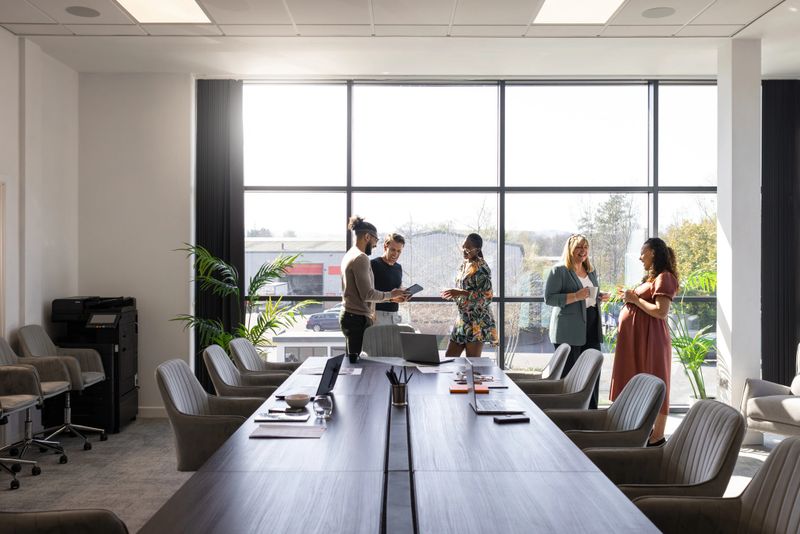 A wide shot of a diverse business meeting being held in a modern conference room. The colleagues are enjoying a coffee break together while standing and talking. Two women are standing, talking while having a cup of coffee. The other colleagues are looking at a digital tablet while talking. Large square windows create a light, positive environment. They are all wearing smart-casual business attire.Videos are available similar to this scenario.