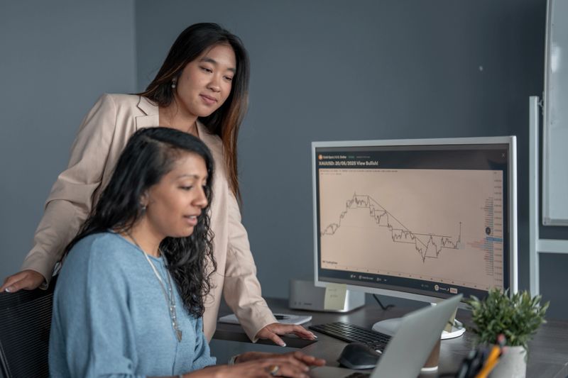 Two professional women discussing financial data displayed on a computer screen in an office. They appear engaged and focused, showcasing teamwork, business analysis, and a professional work environment.