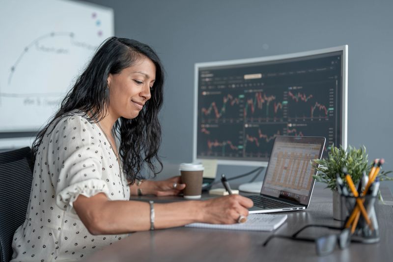 A professional woman attentively works at her desk analyzing financial data and trend charts, showcasing productivity, focus, and engagement within an office environment that promotes modern and organized workspace ethics.