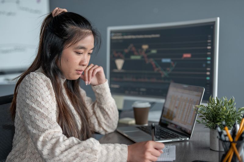 Young woman deeply focused analyzing financial data on a laptop and monitor in an office. Perfect for concepts related to finance, data analytics, stock markets, and professional work environments.