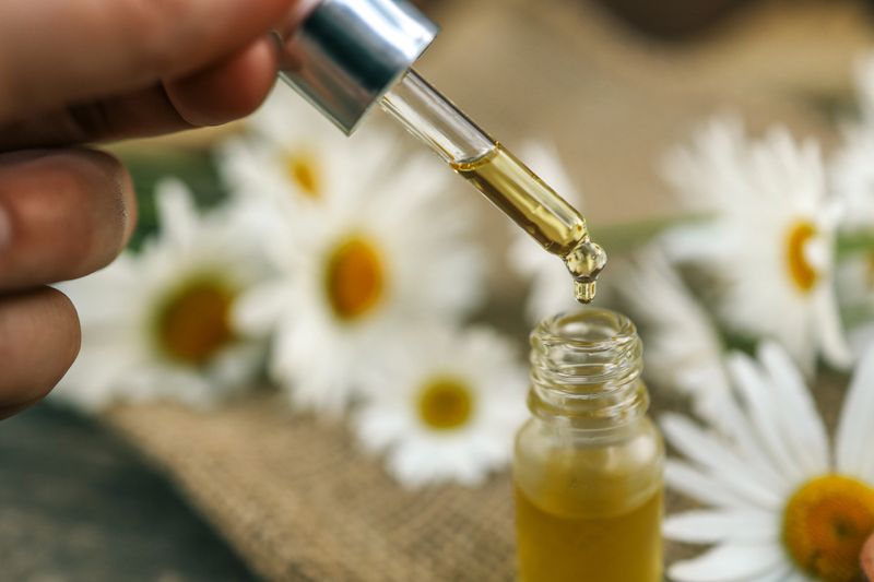 Glass dropper dispensing herbal oil into a small bottle, surrounded by chamomile flowers and wooden spoon on rustic fabric, evoking a natural skincare and wellness theme.