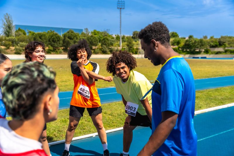 Diverse group of young athletes stretching and warming up on a blue running track with their coach before a race, demonstrating teamwork and dedication