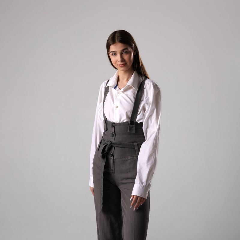 A model displays a fashionable look wearing a white shirt and gray trousers, showcasing elegant style in a minimalist studio setting.