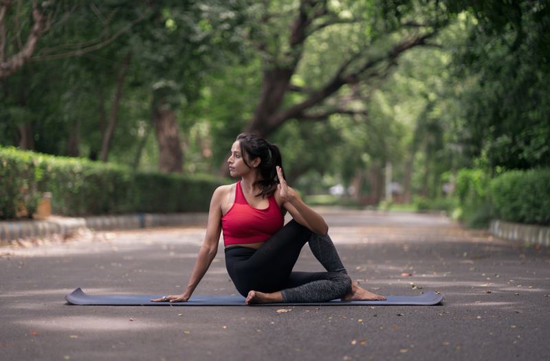 A focused young woman performs the Cow Face Pose (Gomukhasana) on a yoga mat in the middle of a quiet street surrounded by greenery. Dressed in athletic wear, she embraces mindfulness and flexibility in an open-air urban park setting. The image reflects themes of wellness, healthy lifestyle, and self-discipline.