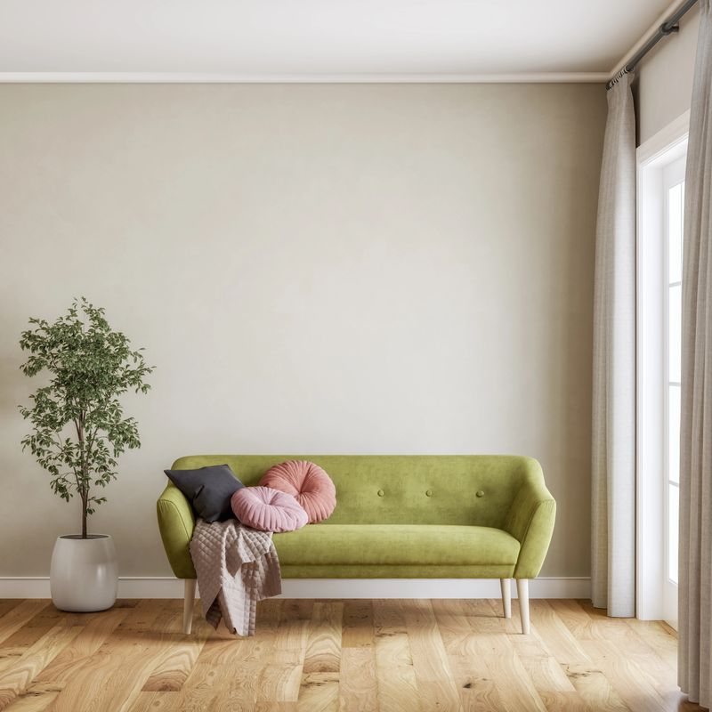 Bright and stylish minimalist living room featuring a green mid-century modern sofa with pastel pillows and a cozy throw blanket. The space is accented by a potted indoor plant (ficus benjamina) and light wooden flooring, with soft natural light streaming in through large windows with neutral curtains. The neutral beige wall provides ample copy space, making the image ideal for design mockups, advertising, or editorial use. Perfect representation of Scandinavian or contemporary interior design trends. 3D rendered image.