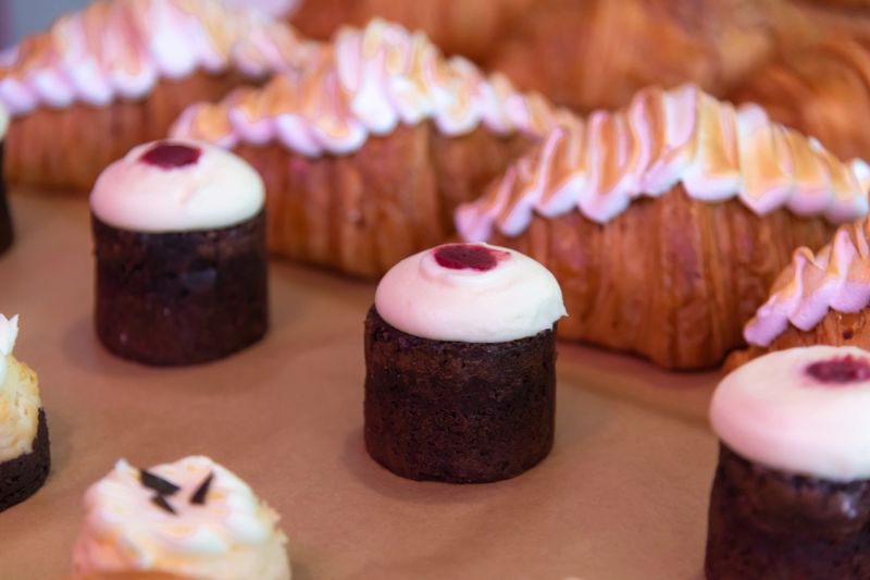 A variety of cupcakes topped with frosting and decorative pastries are arranged neatly on a display table at a bakery. The setting is bright, drawing attention to the treats.