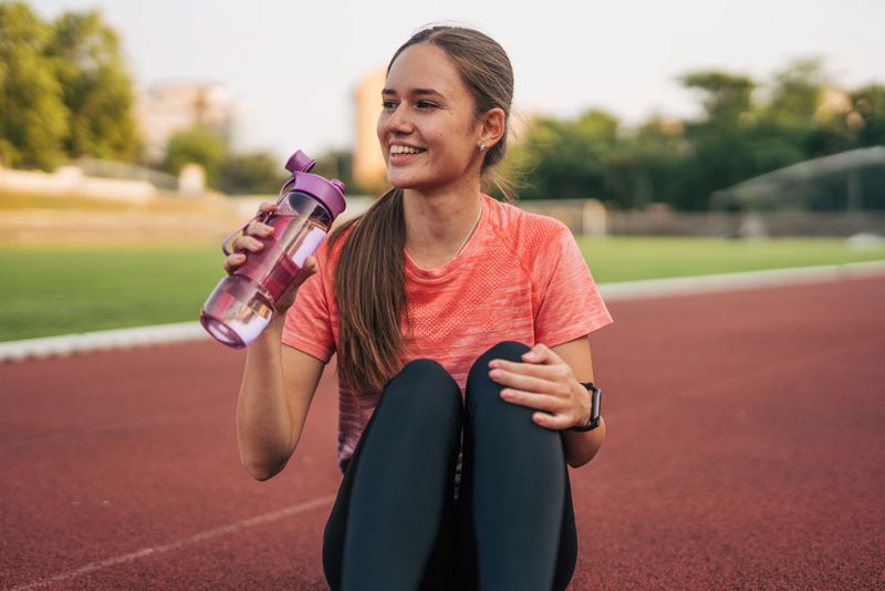 Young female athlete sitting on a running track, enjoying a refreshing drink of water from a bottle while smiling after completing an intense training session