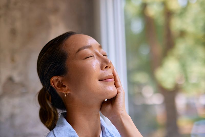 Happy young woman with closed eyes, gently touching her face while enjoying a soothing skincare routine by the window, basking in the warm glow of natural light