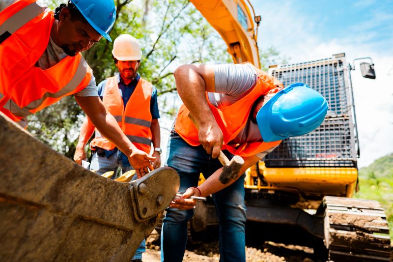 Construction workers examining the bucket of an excavator at an active construction site surrounded by dirt and greenery