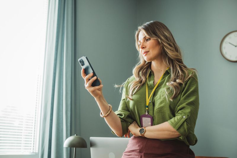 Beautiful middle aged woman working online in office. Leaning on table and using smart phone.